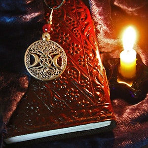 May include: A silver pendant with a triple moon symbol and a pentagram inside a circle, hanging from a red beaded chain, on top of a brown leather journal with intricate floral designs. A lit candle sits on a blue glass holder in the background.
