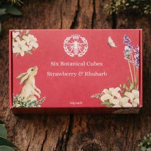 Puede incluir: Una caja roja con el texto "Six Botanical Cubes Strawberry & Rhubarb" e ilustraciones de un conejo, flores y una abeja. La caja está rodeada de follaje verde y descansa sobre una superficie de madera.