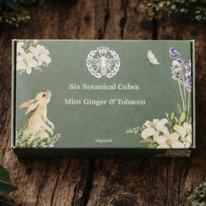 May include: A green box with the text "Six Botanical Cubes" and "Mint Ginger & Tobacco" is surrounded by greenery. The box features a rabbit, flowers, and a bee emblem. The box is on a wooden surface.