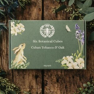 May include: A green box with the text "Six Botanical Cubes" and "Cuban Tobacco & Oak" is surrounded by greenery. The box features a rabbit illustration, floral designs, and a bee emblem. Each cube weighs 10g.