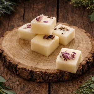 Puede incluir: Una colección de velas de cera cuadradas y claras, cada una adornada con flores secas, se disponen sobre una rodaja de madera rústica. Las velas están rodeadas de vegetación, creando una estética natural y acogedora.