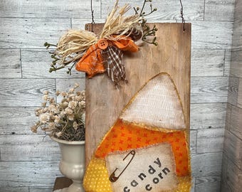 May include: A rustic wooden sign with a fabric candy corn design. The sign is decorated with burlap, a brown and white checkered ribbon, and a safety pin. The sign reads "candy corn".