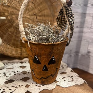 May include: A small orange pumpkin-shaped container with a painted jack-o'-lantern face. The container has a white rope handle and is filled with dried moss. A black and white checkered fabric hangs from the handle.