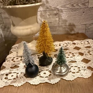 May include: Three miniature Christmas trees with different colored tips. The trees are on a white crocheted doily on a wooden surface.