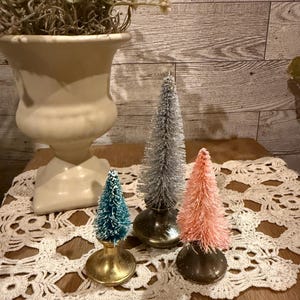 May include: Three miniature bottle brush Christmas trees on a white crocheted doily. The trees are teal, silver, and pink.