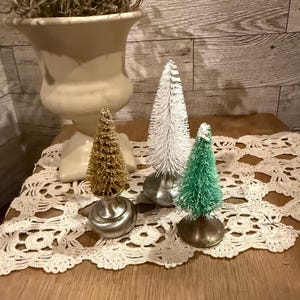 May include: Three miniature bottle brush Christmas trees on a white crocheted doily. The trees are gold, white, and green.
