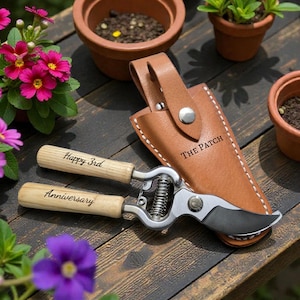 May include: Gardening shears with wooden handles, engraved with "Happy 3rd Anniversary," rest on a wooden surface. A brown leather sheath with "THE PATCH" is next to the shears. Pots of flowers and plants surround the shears.