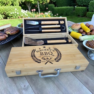Puede incluir: Un juego de herramientas para barbacoa de madera con el texto "MACKEY'S BBQ TOOLS EST 1986" en la tapa. El juego incluye una espátula, un tenedor y unas pinzas con mangos de madera. Las herramientas se muestran en una caja de madera abierta.