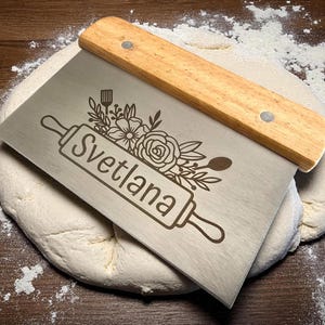 May include: A stainless steel dough scraper with a wooden handle rests on a ball of dough dusted with flour. The scraper is engraved with the name "Svetlana" and floral and kitchen utensil designs.