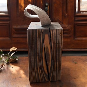 May include: A dark stained wooden doorstop with a natural grain. The block is rectangular with a split down the middle and a woven fabric handle. The doorstop is designed to hold open a door.