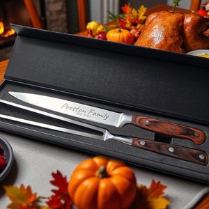 Puede incluir: Un juego de cuchillo y tenedor para trinchar en una caja negra. El cuchillo y el tenedor tienen mangos de madera y hojas de acero inoxidable. El cuchillo está grabado con "Preston Family Est. 2022". El juego está sobre una mesa.