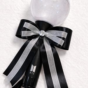 May include: A black light stick with a clear, spherical top and a black handle. A large black bow with silver ribbon accents and a rhinestone center adorns the handle. The BTS logo is visible on the handle.