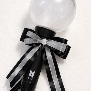 May include: A black and silver light stick with a clear, spherical top. The light stick has a black handle with the letters "BTS" and a logo. A black bow with silver trim and a rhinestone center adorns the base of the sphere.