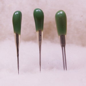May include: Three green and silver sewing needles with pointed tips.