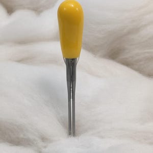 May include: A sewing tool featuring a smooth, rounded yellow handle attached to a slender, two-pronged metal fork. The metal prongs are straight and parallel, designed for precision work, and the handle provides a comfortable grip for the user.