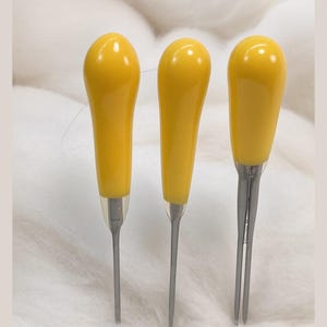 May include: Three sewing tools with smooth, bright yellow teardrop-shaped handles and metallic silver tips. Two of the tools feature a single pointed needle end, while the third tool on the right has a two-pronged fork end for precision work.