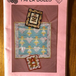 May include: A pink paper with the text "PAPER DOLLS" and illustrations of paper doll quilts. The main quilt is light blue with heart and doll designs. Two smaller framed doll images are also shown.