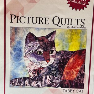 May include: A colorful patchwork quilt pattern featuring a tabby cat. The quilt is made of various fabrics in shades of blue, red, orange, and yellow. The quilt is titled "Tabby Cat" and is part of a collection of picture quilts by Sharon Malec.