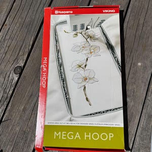 May include: A Husqvarna Viking Mega Hoop embroidery frame in its original packaging. The box features a white background with a graphic of white orchid flowers and the text Mega Hoop, Sewing Area 14.2 x 6, 360 x 150 mm for Designer Series.