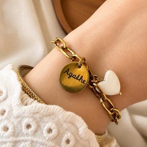 Puede incluir: Pulsera de cadena dorada con un dije redondo grabado con el nombre "Agathe" y un dije en forma de corazón blanco. La pulsera se lleva en la muñeca, con una manga bordada blanca en el fondo.