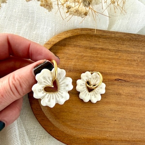 May include: White flower-shaped earrings with a gold heart-shaped center and gold hoops. The earrings are displayed on a wooden tray. The earrings are handmade and have a textured surface.