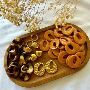 May include: A wooden tray showcases an assortment of jewellery. There are gold-toned earrings, heart-shaped pendants, and donut-shaped earrings in brown and gold hues. Dried flowers are arranged above the tray.