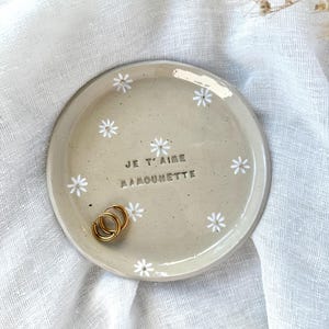 以下が含まれることがあります： 中央に「JE T'AIME MAMOUNETTE」という暗い文字が刻印された、ベージュ色の丸いセラミック製トレイ。手描きの白いデイジーのモチーフと金色の中心部が特徴です。表面には小さな金色のフープピアスが2つ置かれています。