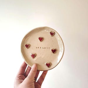 以下が含まれることがあります： 斑点のある釉薬をかけた小さな白い陶器の皿。皿の縁には赤いハートが6つあり、中央には「mon amour」という文字が刻まれている。