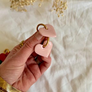 Könnte beinhalten: Ein Paar hellrosa herzförmige Ohrringe mit goldfarbenen Reifen. Die Ohrringe haben eine glänzende, einfarbige Oberfläche und werden von einer Hand gehalten. Der Hintergrund ist eine neutrale Farbe.