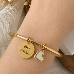Puede incluir: Pulsera dorada con dos colgantes. Uno es un disco redondo con los nombres "Ezio Milena" grabados. El otro es un colgante en forma de corazón con relleno de esmalte blanco. La pulsera se lleva en la muñeca.