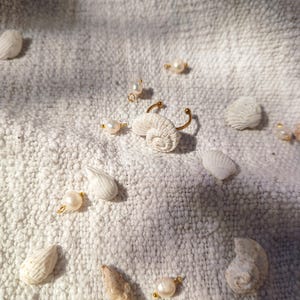 Puede incluir: Conchas blancas y perlas esparcidas sobre un fondo de tela blanca texturizada. Un anillo de oro con un diseño de concha blanca está en el centro de la imagen.