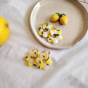 Puede incluir: Un par de pendientes de aro dorados con un dije blanco en forma de flor. El dije está decorado con un estampado de limón amarillo y hojas verdes.