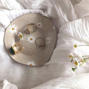 May include: A small, round, beige ceramic dish with a textured surface and white daisy designs. The dish is filled with gold rings and a ring with a green stone.