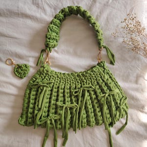 May include: A green crocheted handbag with a long strap and multiple bows. The bag has a small matching heart-shaped keychain.