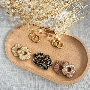 May include: A collection of jewelry on a wooden tray, including two pairs of plain gold hoop earrings and three pairs of flower-shaped earrings in cream, black, and dusty pink, each featuring gold-toned radial line detailing on the petals.