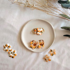 May include: A set of earrings featuring a white flower design with orange and green accents. The earrings are displayed on a white plate with a white background.