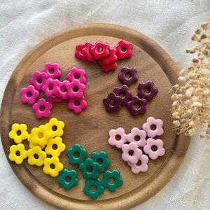 Puede incluir: Una bandeja de madera exhibe una variedad de cuentas coloridas en forma de flor. Las cuentas son en tonos de rojo, rosa, amarillo, verde y morado. Las cuentas están dispuestas en pequeños grupos en la bandeja.