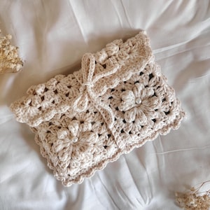Könnte beinhalten: Eine beige gehäkelte Clutch mit floralem Design. Die Clutch ist aus weichem, strukturiertem Garn gefertigt und hat einen gewellten Rand. Ein passender Kordelverschluss sichert die Oberseite. Die Clutch liegt auf einer weißen Oberfläche.