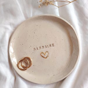 May include: A speckled, off-white ceramic trinket dish with the word "MARRAINE" embossed on the surface. Two gold hoop earrings and a gold heart-shaped charm rest on the dish. The dish is round with a slightly raised edge.