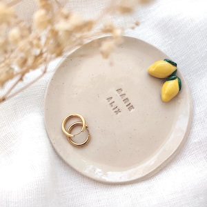 Puede incluir: Un plato pequeño de cerámica blanquecina moteada con el texto en relieve "MARIE ALIX". Dos pendientes de aro dorados y dos adornos de cerámica amarillos con forma de mango están sobre el plato. El plato está sobre una superficie blanca.