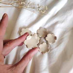 Pode incluir: Cinco pequenos pratos de cerâmica em forma de nuvem, na cor bege creme com detalhes salpicados. Os pratos estão dispostos sobre uma superfície de tecido branco, com uma mão segurando um dos pratos. Flores secas ao fundo.