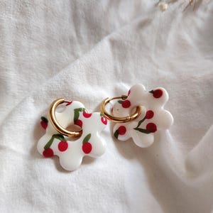 Puede incluir: Un par de pendientes blancos con forma de flor con detalles de cerezas rojas y aros dorados. Los pendientes están sobre un fondo de tela blanca, con unas ramitas de flores secas en la esquina superior derecha.