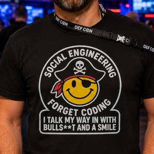 May include: A black t-shirt featuring a yellow smiley face wearing a pirate hat with a skull and crossbones and an eye patch. The text reads: SOCIAL ENGINEERING FORGET CODING I TALK MY WAY IN WITH BULLS**T AND A SMILE. A black DEF CON lanyard is worn.