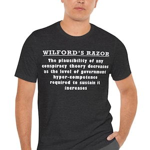以下が含まれることがあります： 「WILFORD'S RAZOR」の文字と陰謀論に関する記述が白でプリントされた濃いグレーのTシャツ。テキストは中央に配置されています。Tシャツは半袖です。