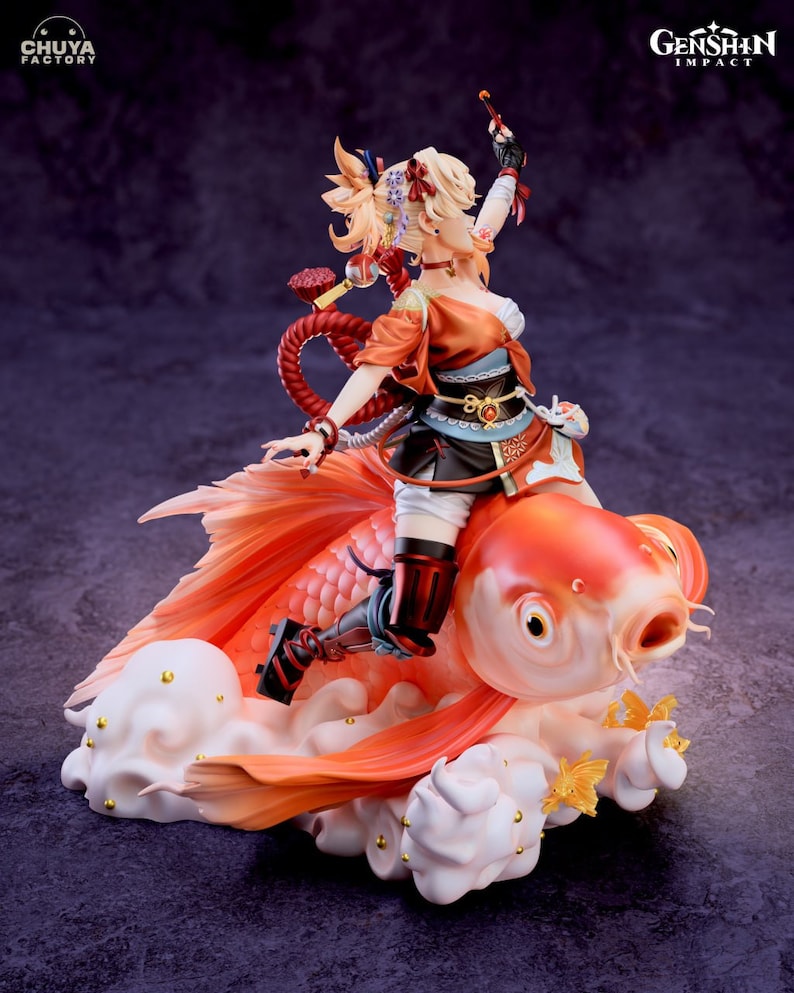 May include: A figurine of a female character from the video game Genshin Impact, riding a large, orange goldfish. The character is wearing a red and black outfit with a long, flowing red scarf. The goldfish is sitting on a white cloud with gold accents. The figurine is made of high-quality plastic and is very detailed.