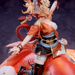 May include: A figurine of a female character from the video game Genshin Impact. She is wearing a red and orange outfit with a white sash and a black belt. She is riding a large, orange goldfish. The figurine is made of high-quality plastic and is very detailed.