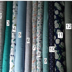 May include: A collection of various fabric rolls, showcasing diverse patterns and colors. The fabrics include floral prints, dinosaur motifs, and nautical designs. Colors range from soft pastels to deep blues, with some featuring stripes and animal prints. The rolls are tightly packed together.