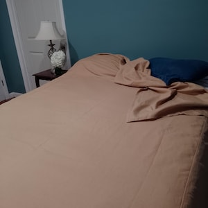 May include: A bed with a tan sheet set and a blue pillow. The bed is made, with the top sheet partially pulled back. A lamp and a small table are visible to the left, and a decorative wreath hangs on the teal wall.