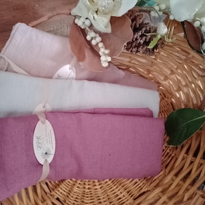 May include: Rolled linen napkins in shades of pink and white, displayed in a woven basket. The napkins are tied with ribbons and tags. Decorative floral accents and a pinecone add to the presentation. The napkins are a soft, natural fabric.