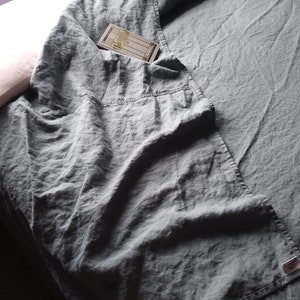 May include: A crumpled, slate-blue linen sheet covers a bed, with a pillow visible in the upper left corner. A small tag is visible on the sheet. A folded section of the sheet has a pocket with a small card inside.